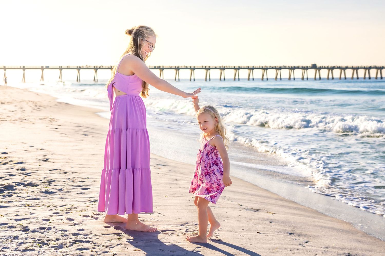 Two people in purple dresses enjoy a sunny day walking along the beach near a fishing pier.