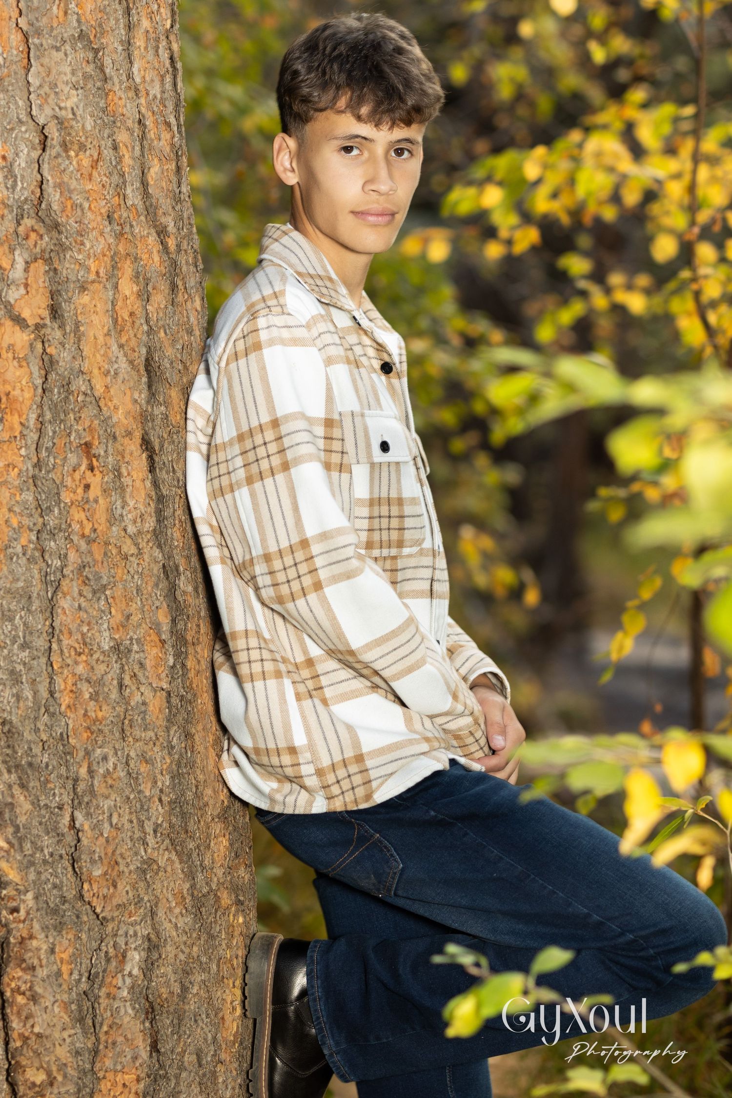 Portrait of person leaning against tree trunk wearing plaid flannel shirt.