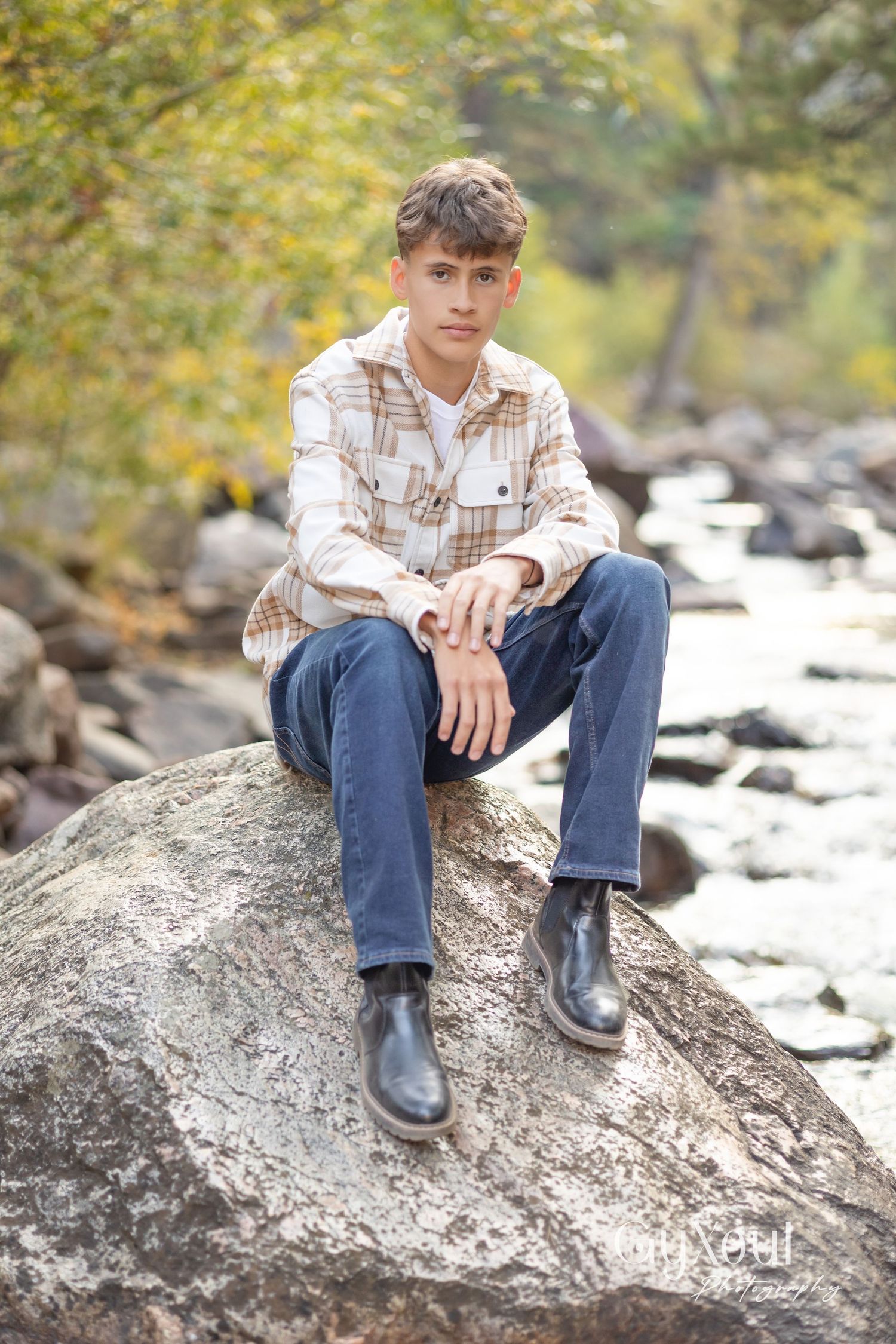 Person in plaid shirt sits on large rock by stream wearing dark boots.