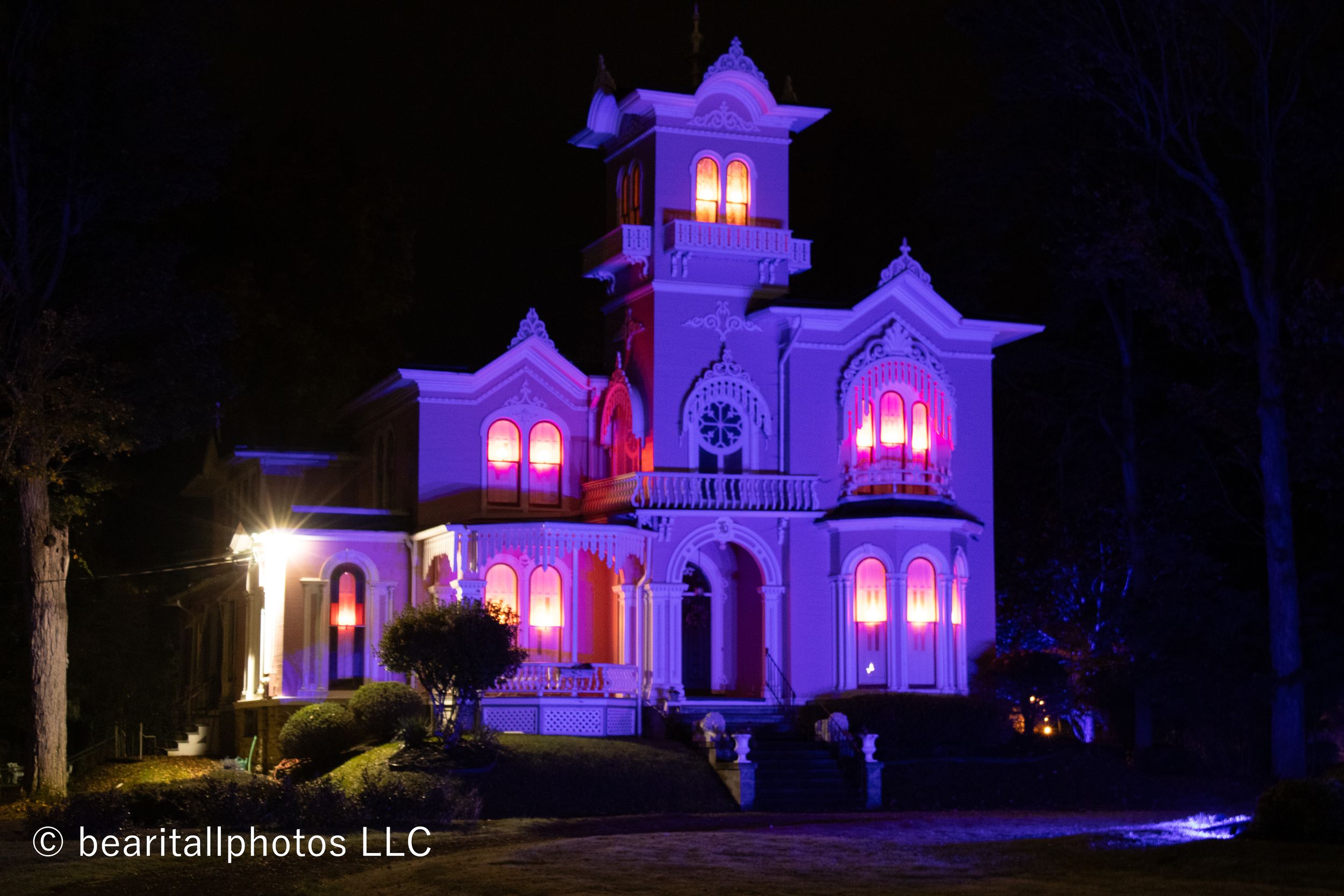 Victorian mansion illuminated in purple and pink lighting against dark night sky with glowing windows.