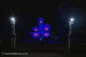Wide angle shot of blue-lit mansion with bright spotlights and ornate fence posts.