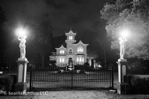 Black and white panoramic view of illuminated mansion with statues along fence at night.