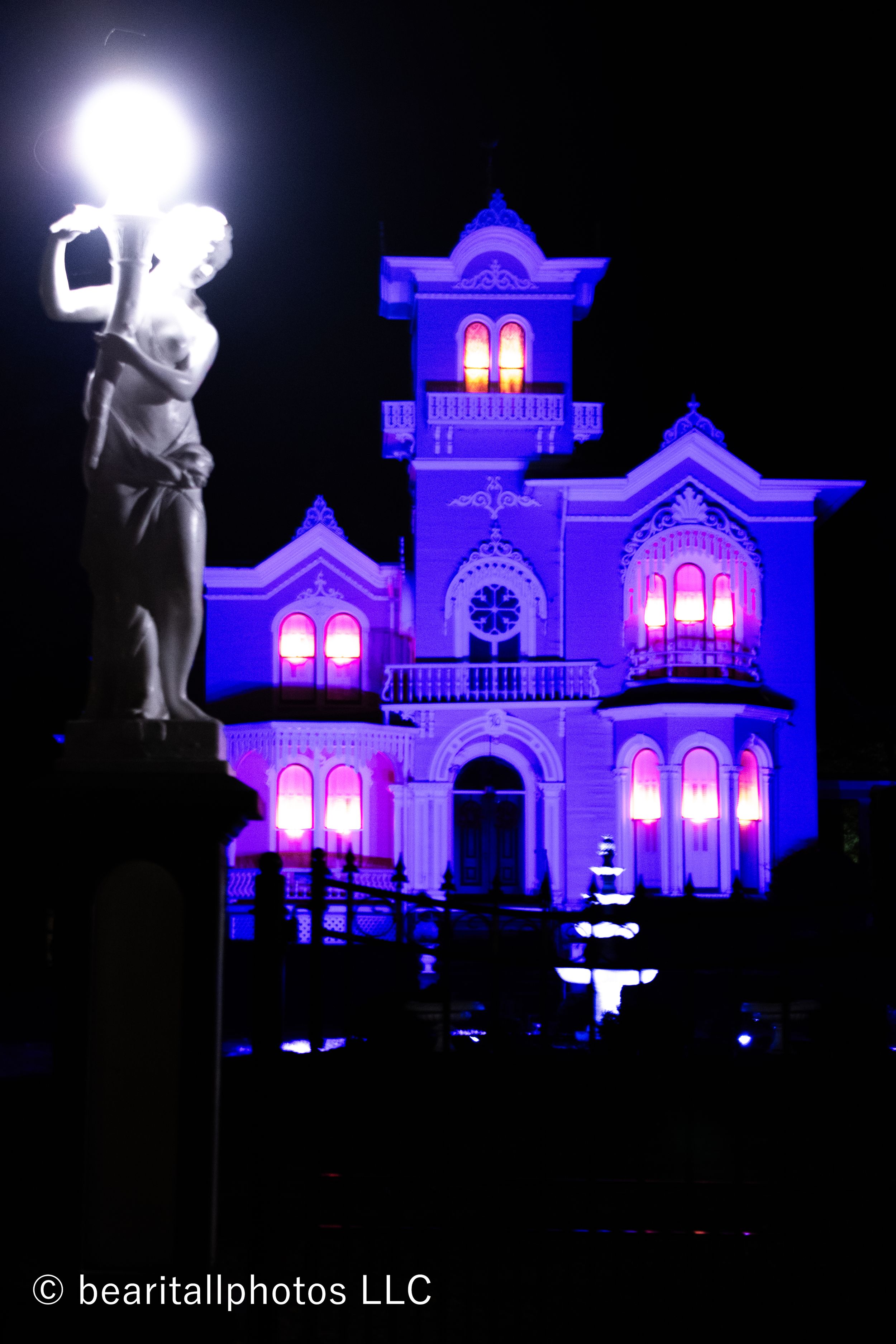 Gothic revival mansion illuminated in blue with classical statues silhouetted in foreground.