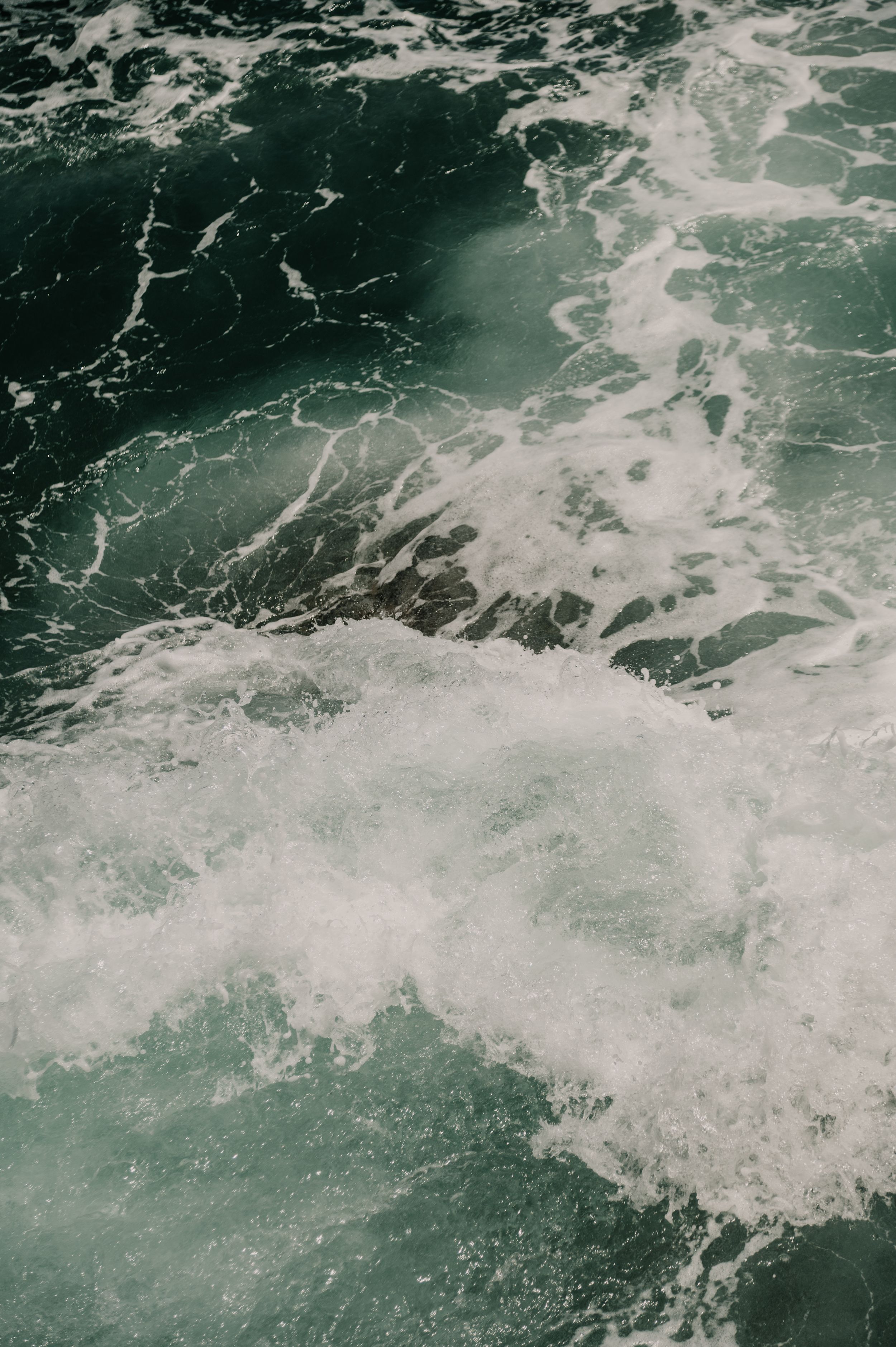 Churning ocean waves create a dramatic pattern of white foam on dark turquoise water.