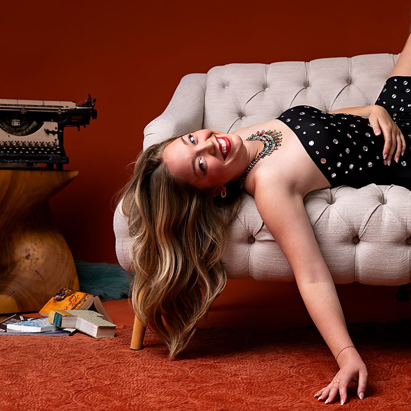 A person in a polka dot dress reclines on a beige tufted sofa against a rust-colored wall.