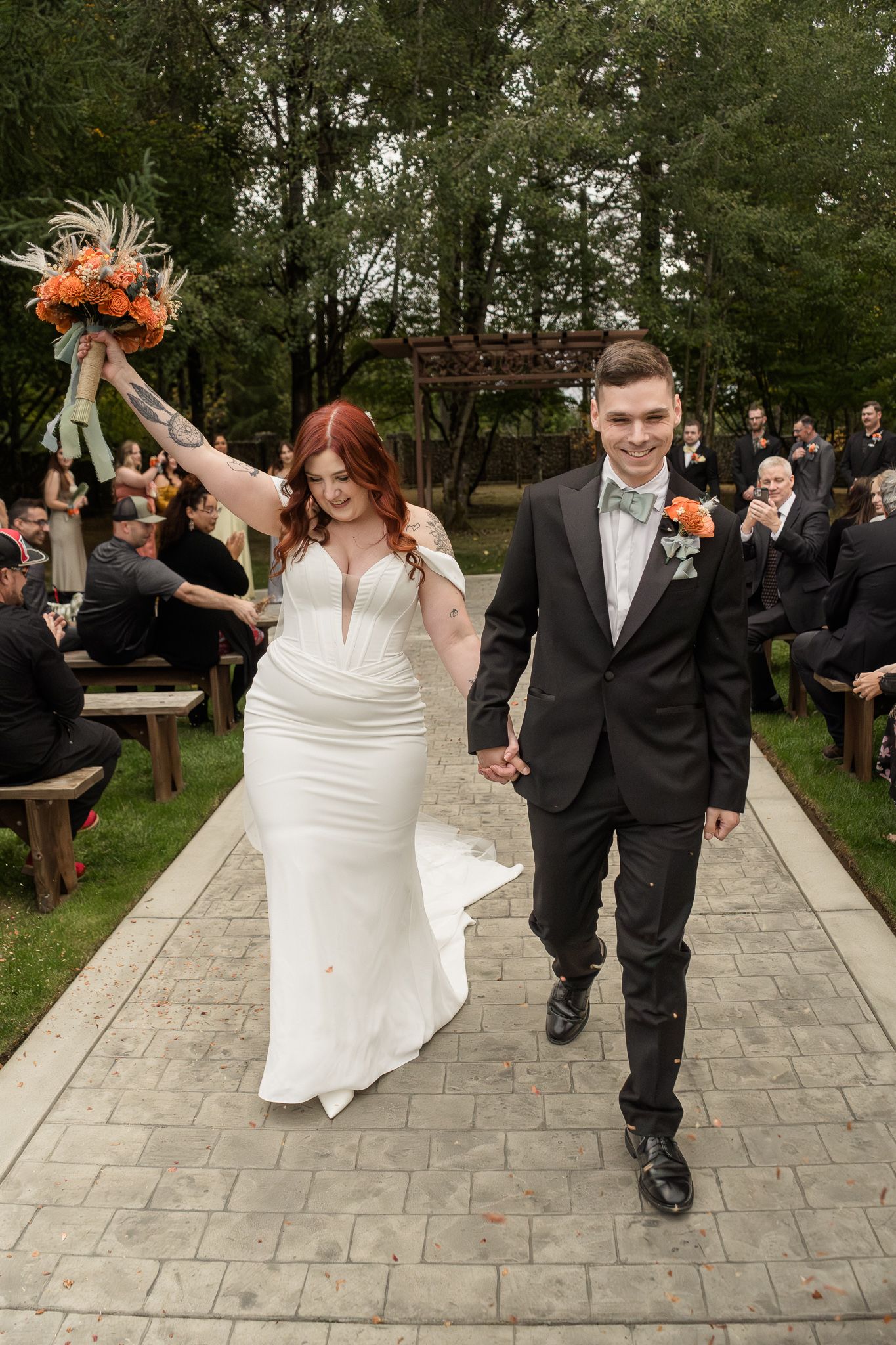 Wedding couple celebrates on stone pathway with guests cheering in outdoor garden setting.