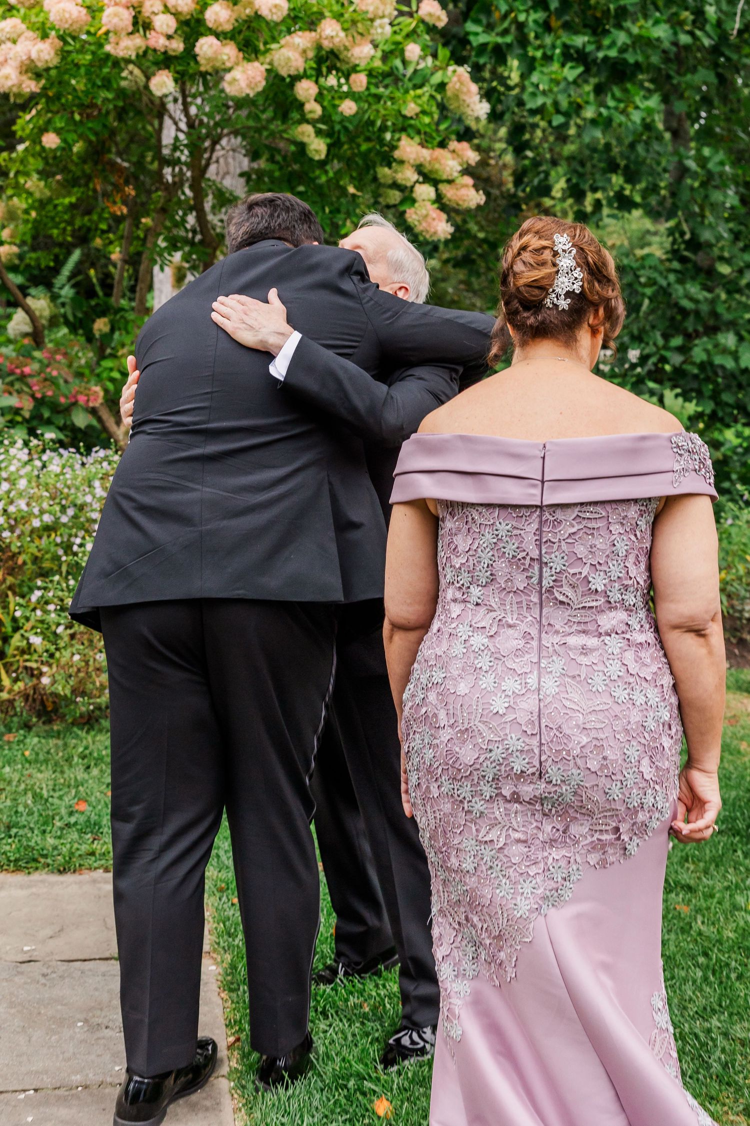 A couple embraces while a woman in a dusty pink formal gown stands nearby in a garden setting with blooming flowers.