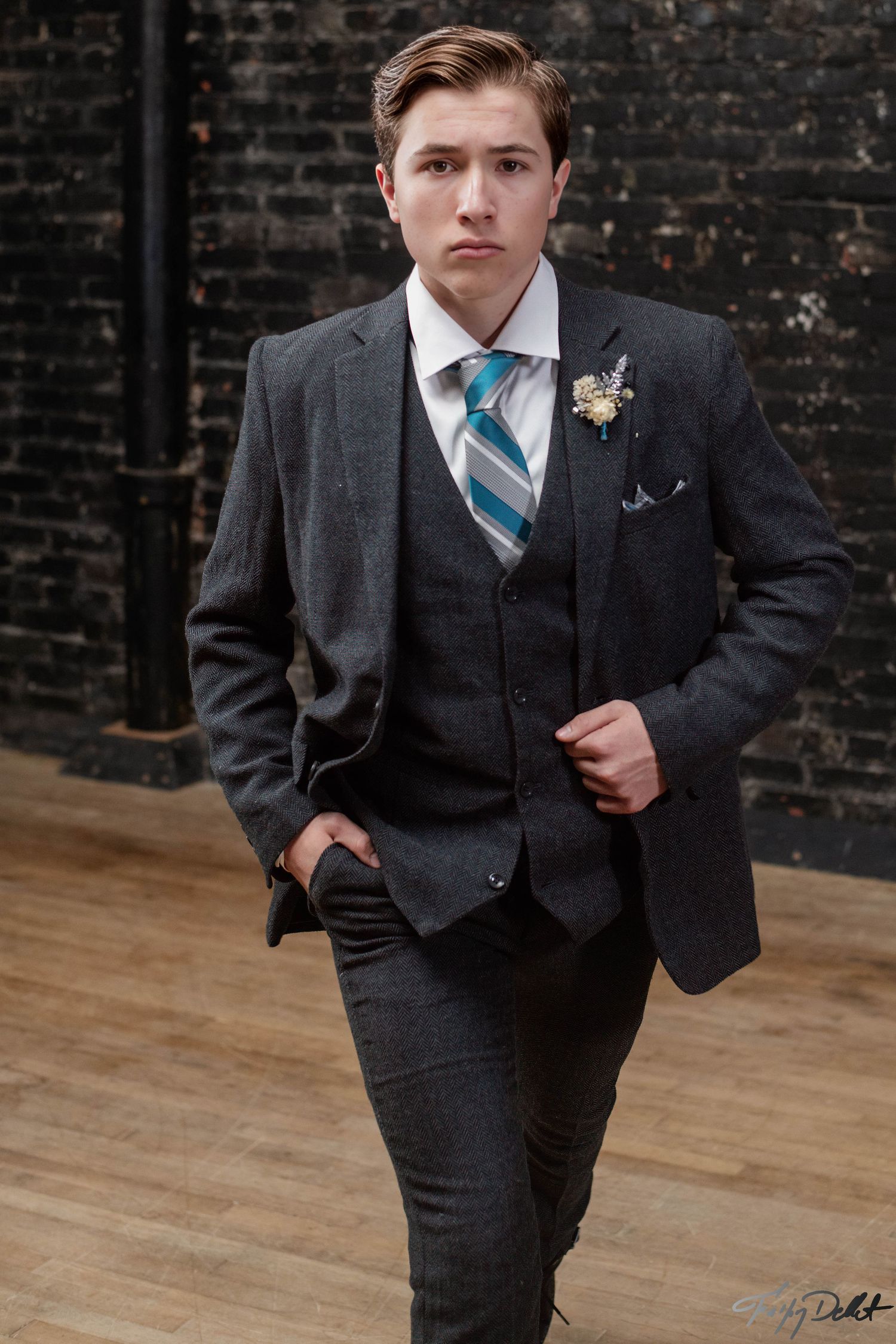 Young boy in dark formal suit with blue striped tie poses against brick wall background.