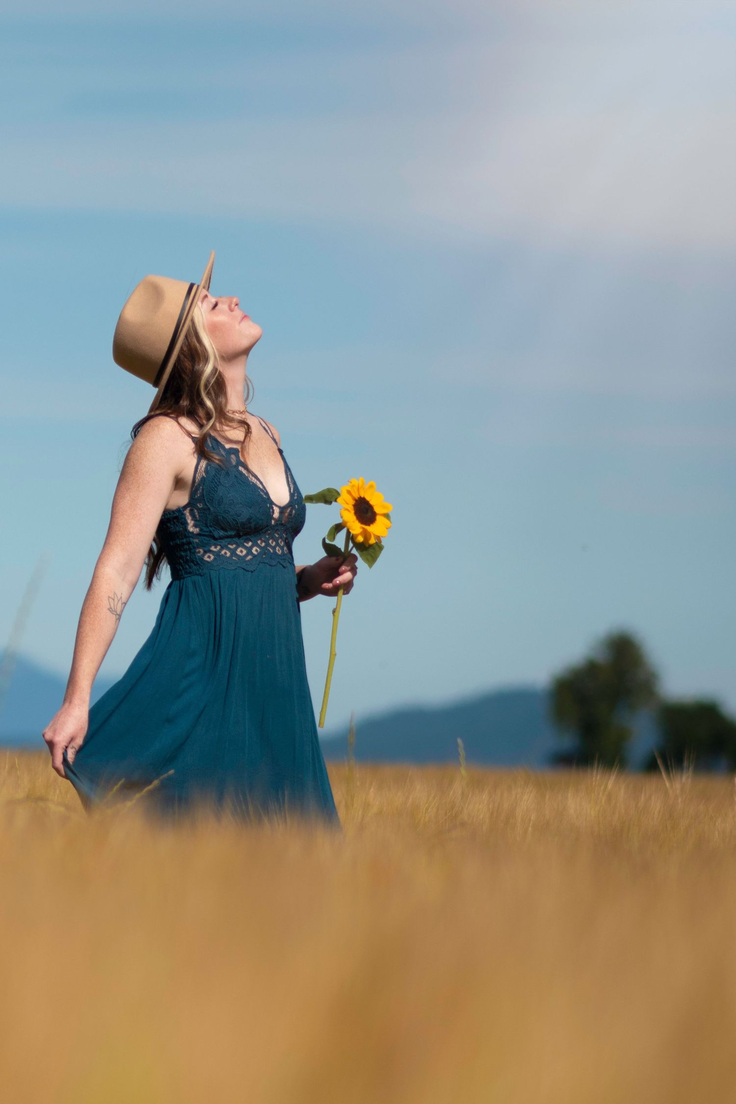 Someone in a blue dress holds a sunflower while standing in a golden wheat field on a sunny summer day.
