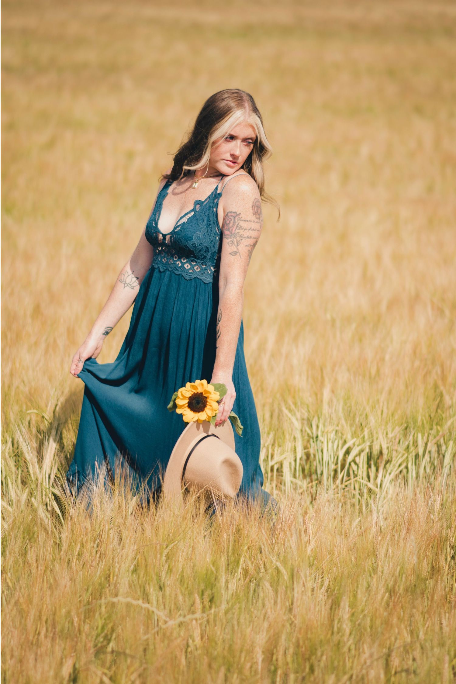 A person in a teal dress poses gracefully in a golden wheat field holding a sunflower during sunset.