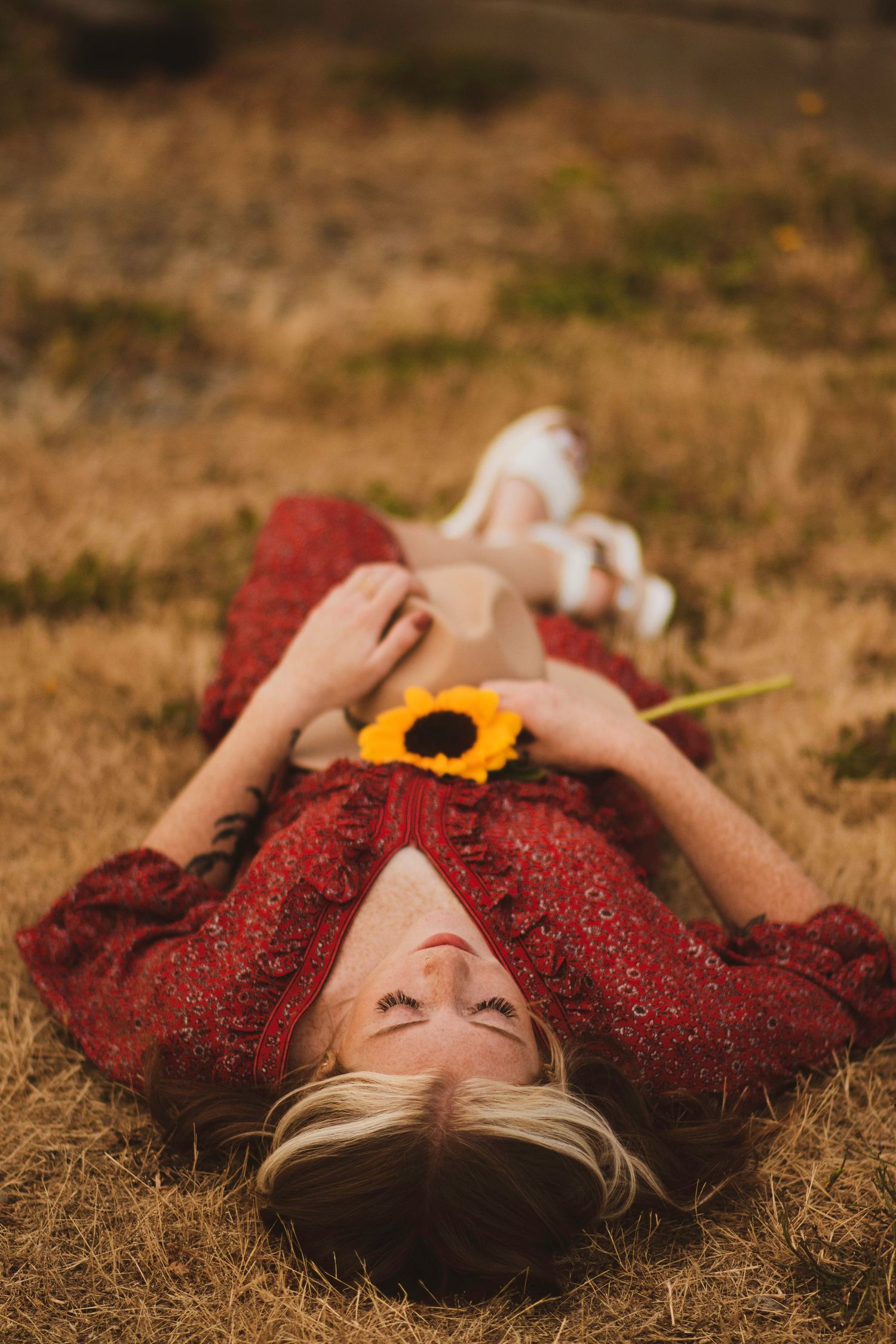 Person in red dress lying on dry grass with yellow sunflower in artistic outdoor portrait series.