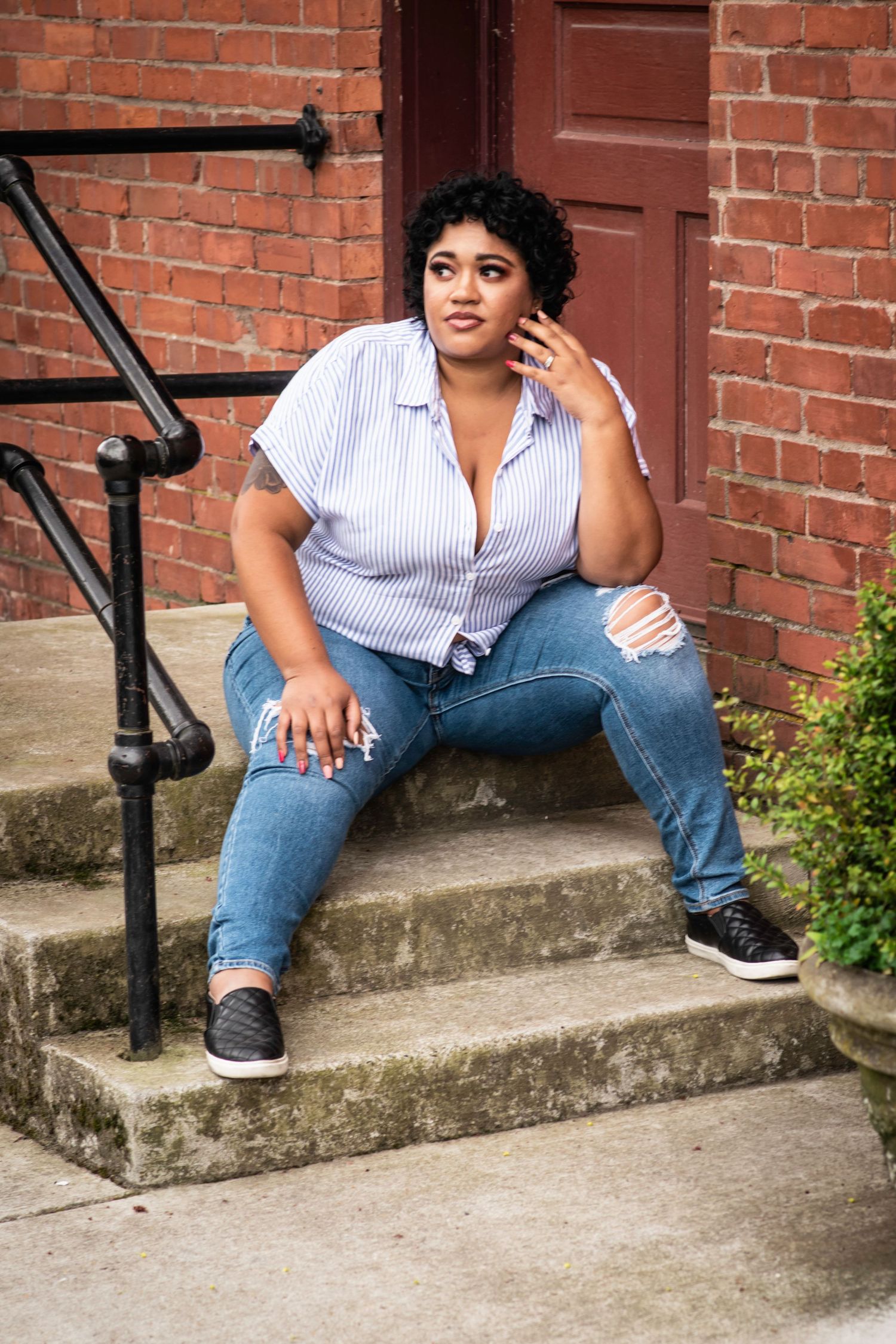 Person in striped shirt and jeans sits casually on concrete steps against red brick wall.
