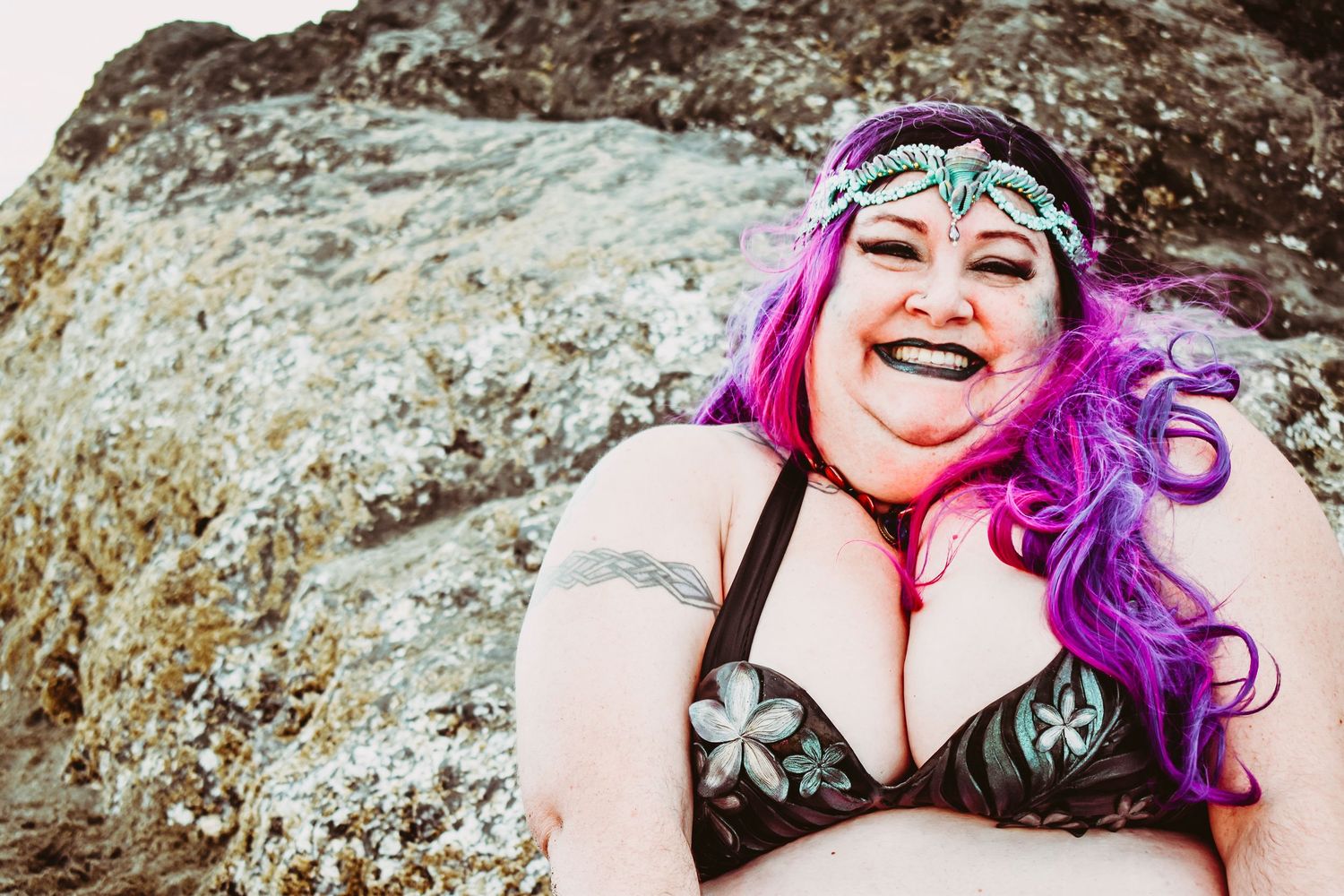 A person with vibrant purple hair poses against rocky terrain in a boudoir photoshoot outdoors.