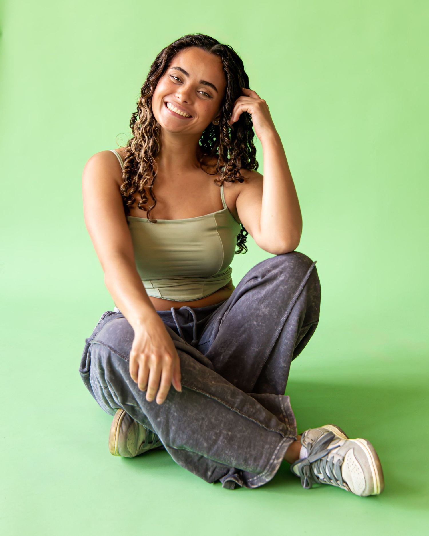 Someone in casual attire sits cross-legged on the floor against a bright green wall, showing relaxed and playful poses.