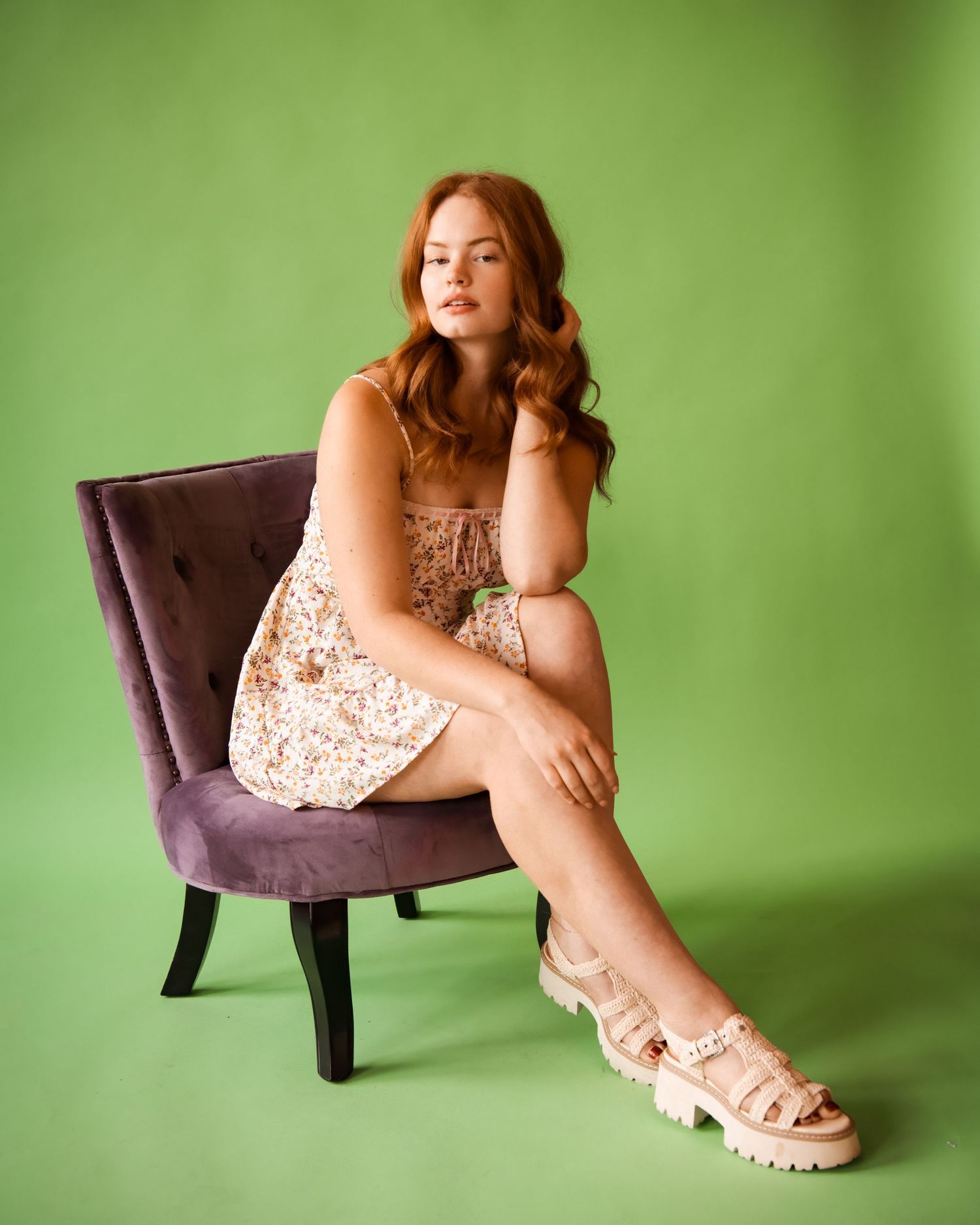 A person in a floral dress poses artistically on a purple chair against a green studio backdrop.