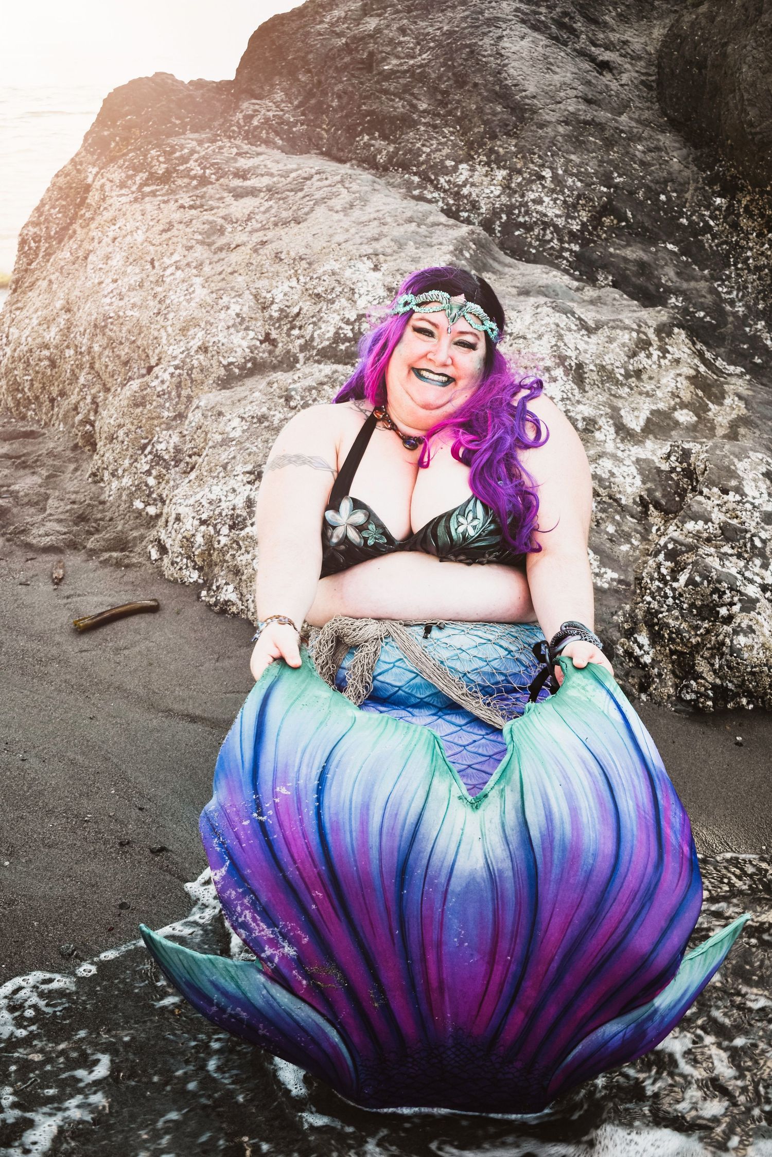 A person wearing a vibrant mermaid costume with purple and blue tail poses playfully on rocky terrain.