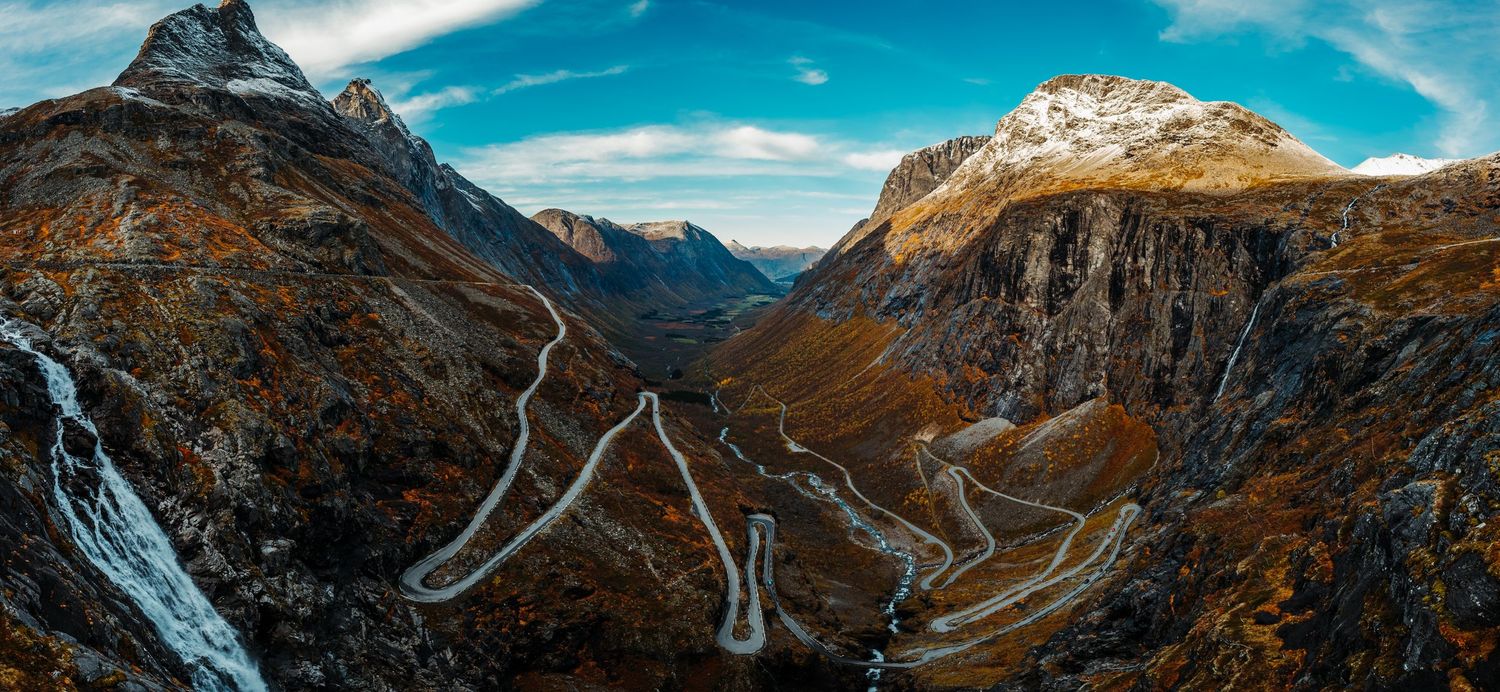 Panoramic mountain landscape with dramatic peaks and winding road under blue sky.