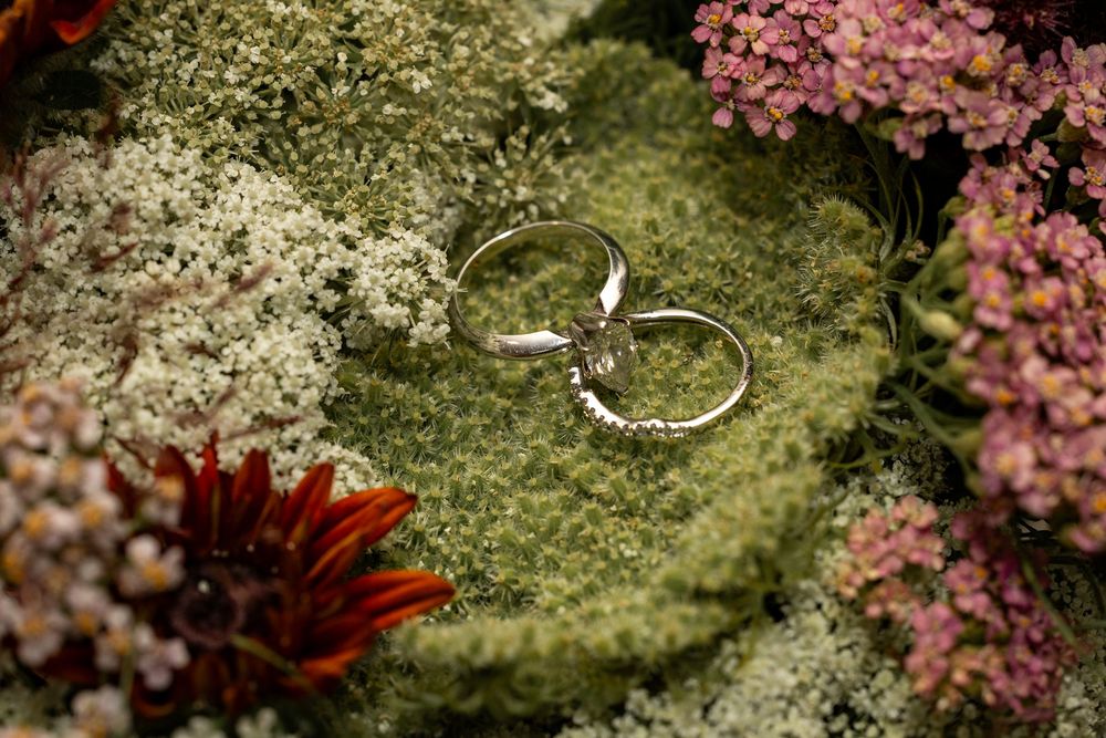 Wedding rings rest on delicate white lace with pink blossoms and greenery creating a romantic backdrop.