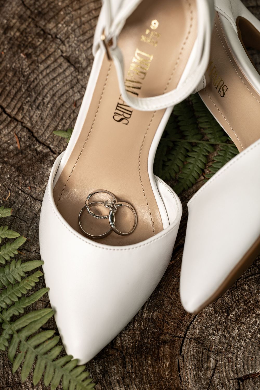 White bridal shoes with wedding rings displayed on rustic wood surface with delicate fern leaves.