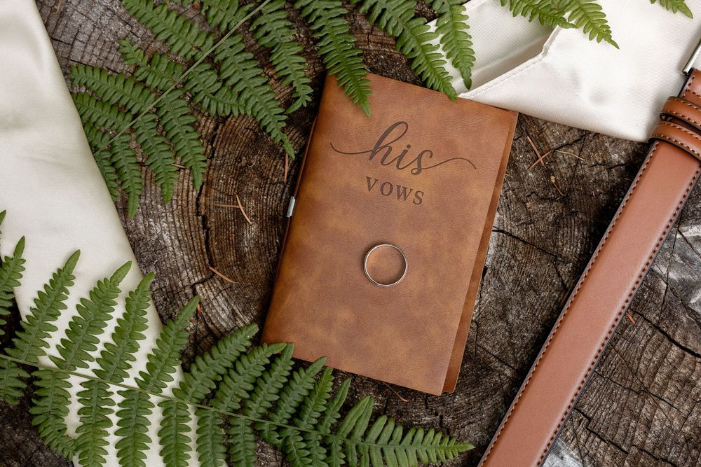 Brown leather journal with strap lies on rustic wood surface next to delicate green fern leaves.