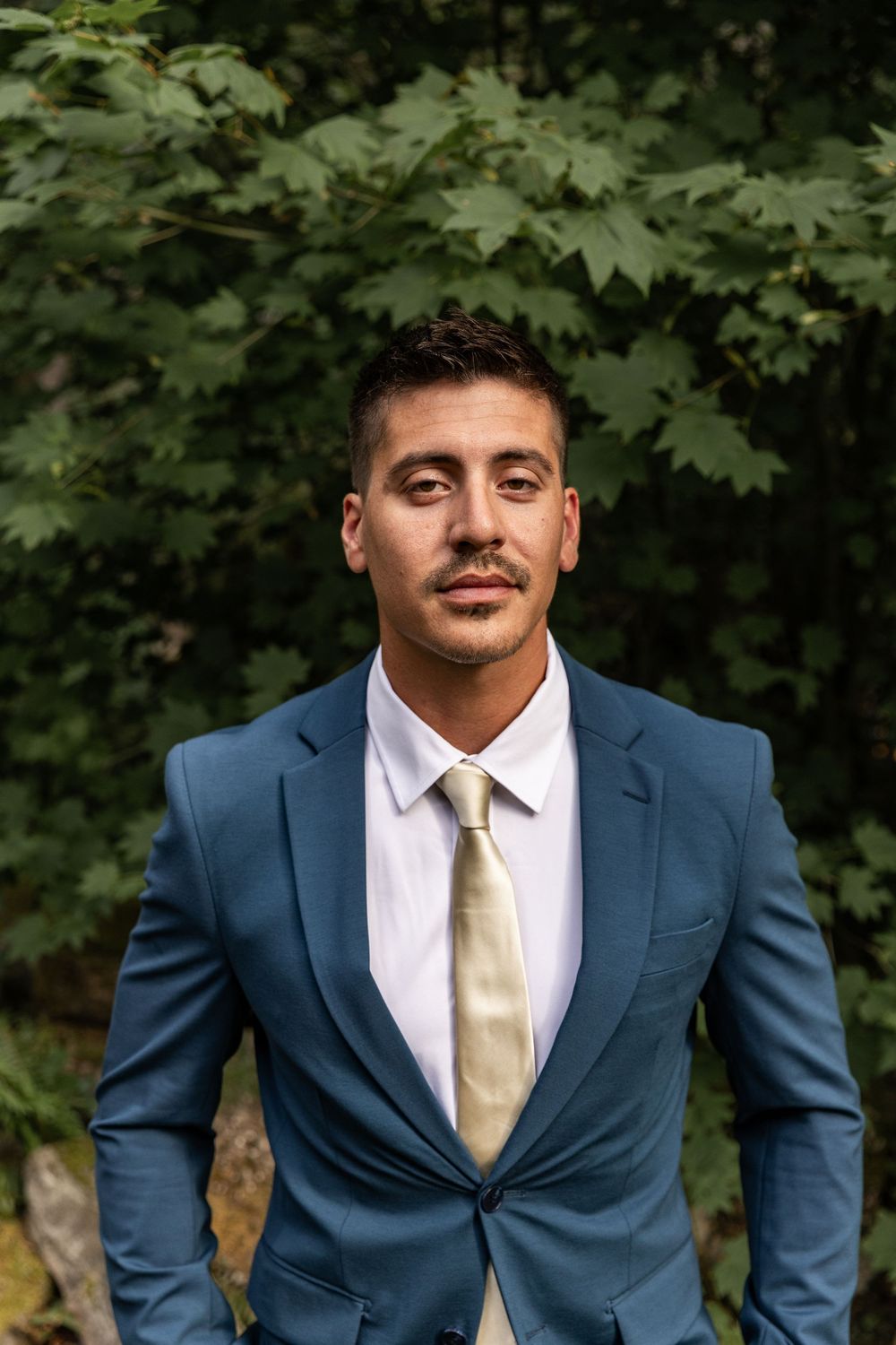 A groom in a navy blue suit and champagne tie poses against a backdrop of green maple leaves.