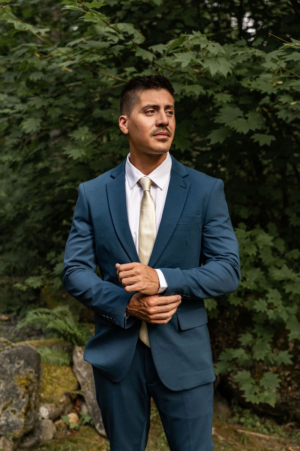 A groom adjusts his navy suit jacket while standing among green foliage.