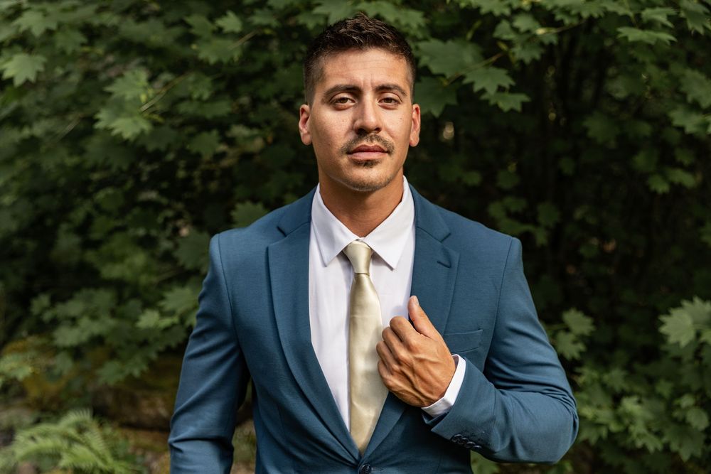 A man in a navy suit adjusts his tie while standing outdoors with green foliage in the background.