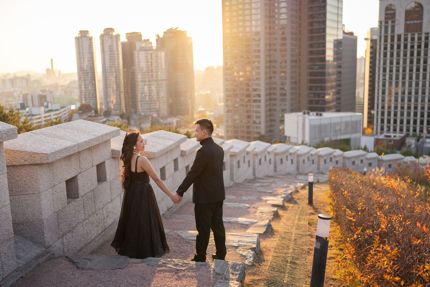 Two people stand on a rooftop terrace overlooking a cityscape at golden hour with tall buildings in the background.