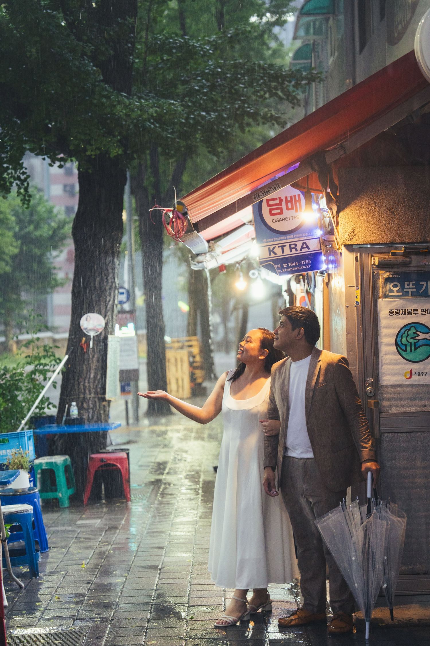 Couple stands outside storefront on rainy evening in urban setting with glowing lights and wet pavement.