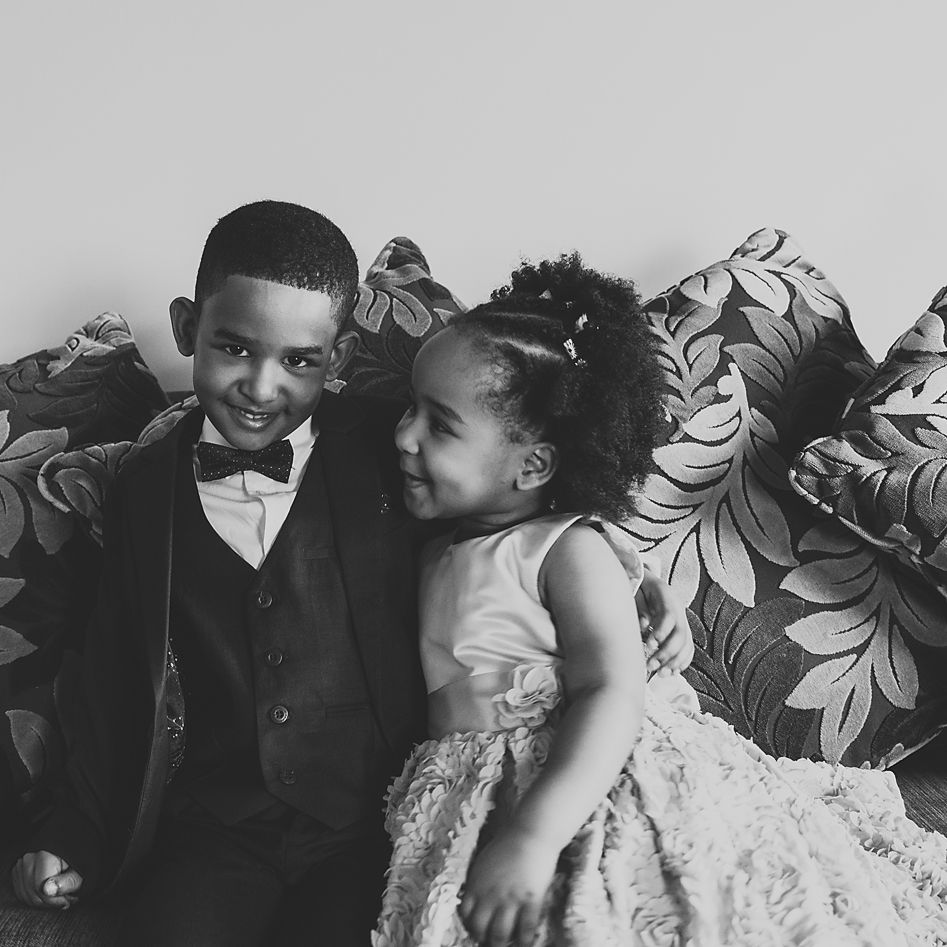 A black and white portrait of two people sitting on a floral couch, one in a tuxedo and one in a dress.