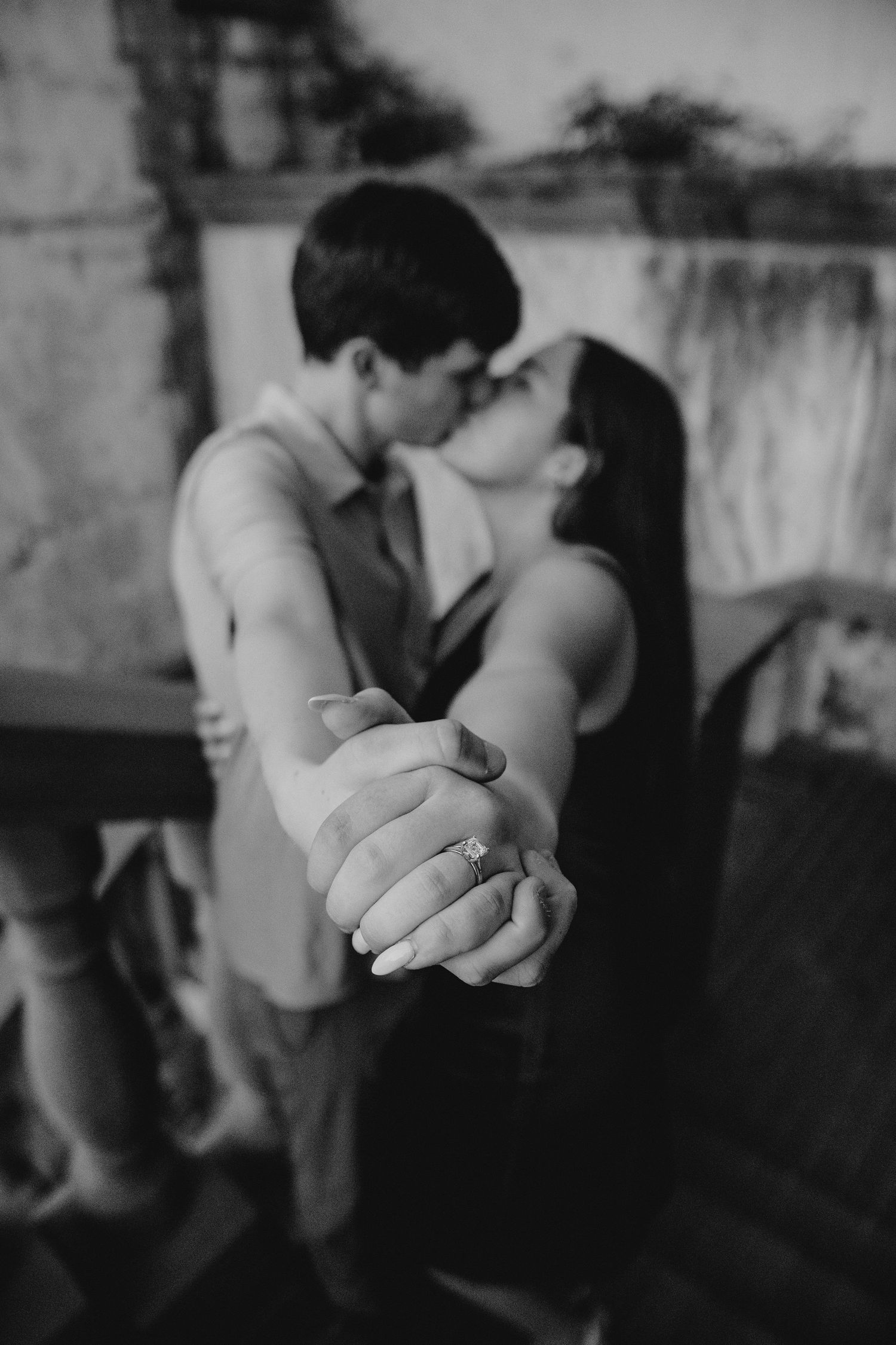 Black and white photo of couple kissing while showing off engagement ring close to camera.