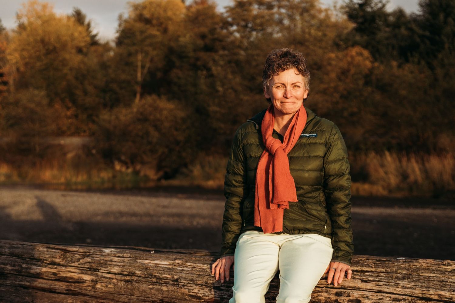 A figure in a red scarf and white pants sits on a rock during golden autumn sunset lighting.