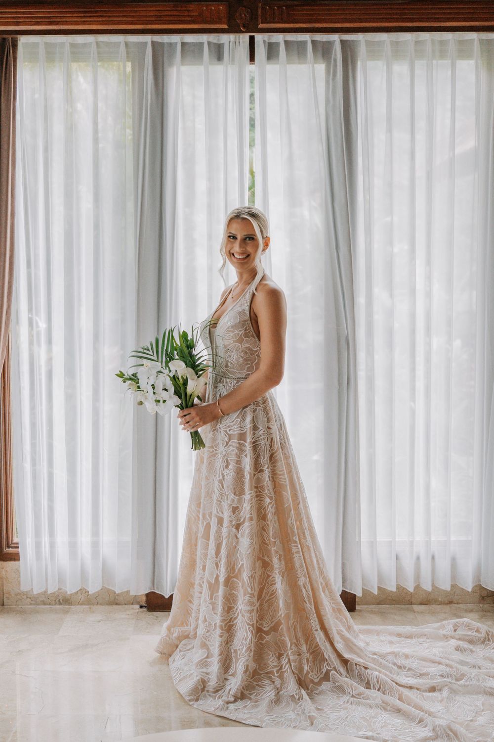 An elegant lace wedding gown with a flowing train is shown in front of sheer white curtains with a floral bouquet.