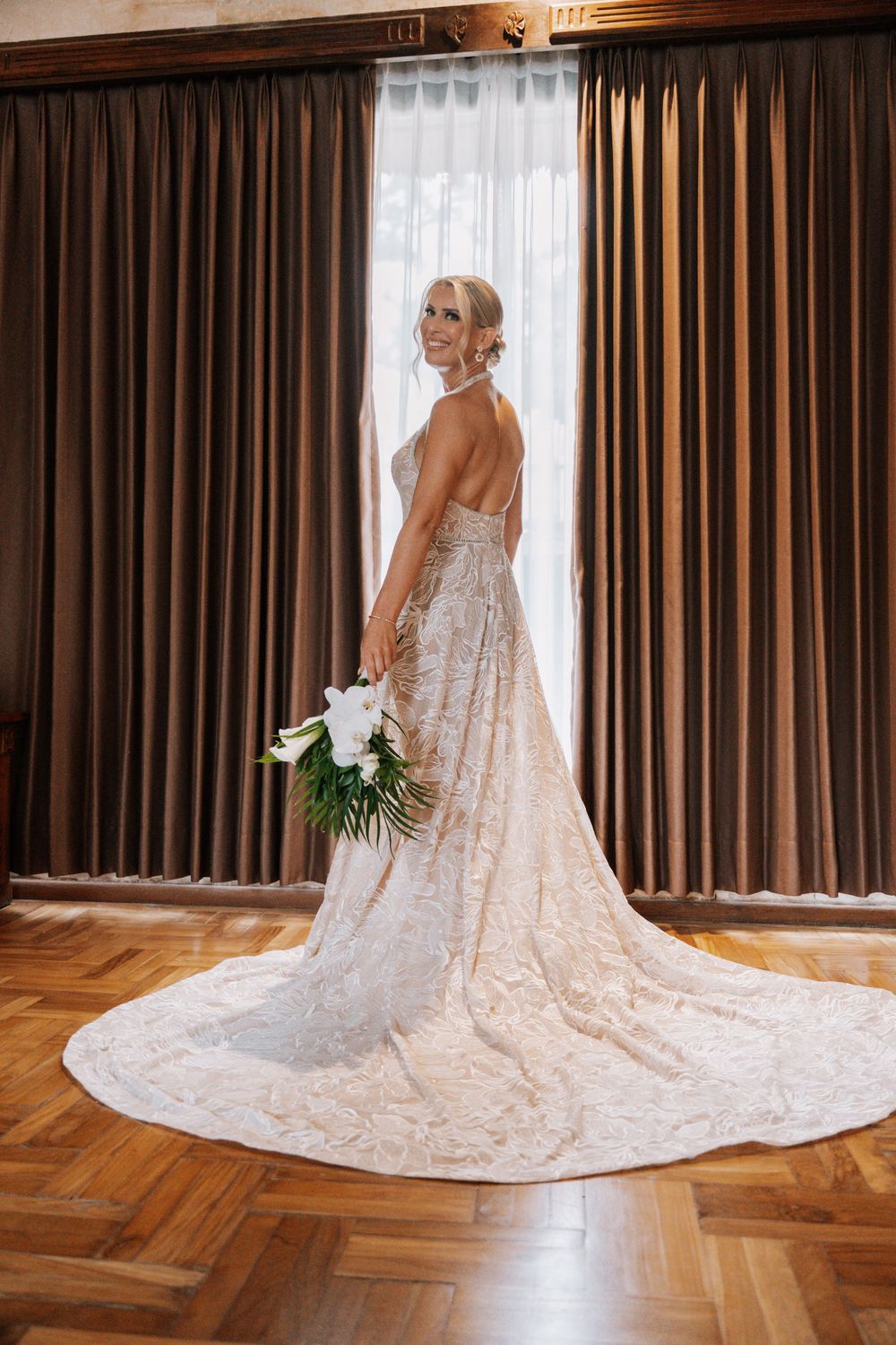 A bride in an elegant white lace wedding gown with a dramatic cathedral-length train stands in a room with wooden floors.