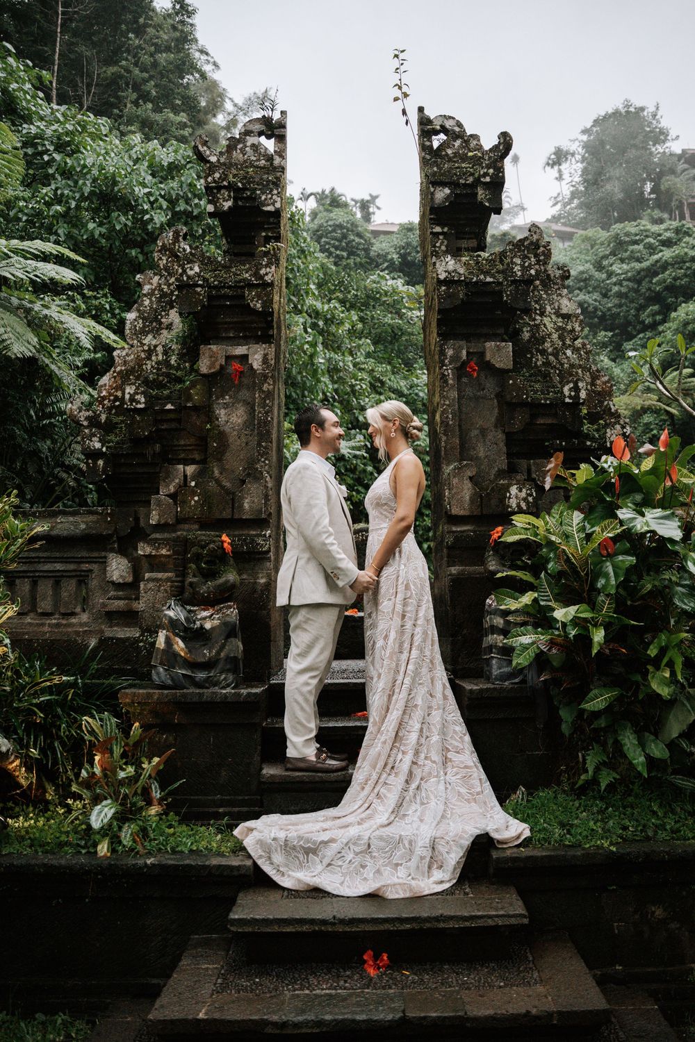 A couple shares an intimate moment in traditional wedding attire at an ancient stone temple in a lush tropical garden setting.