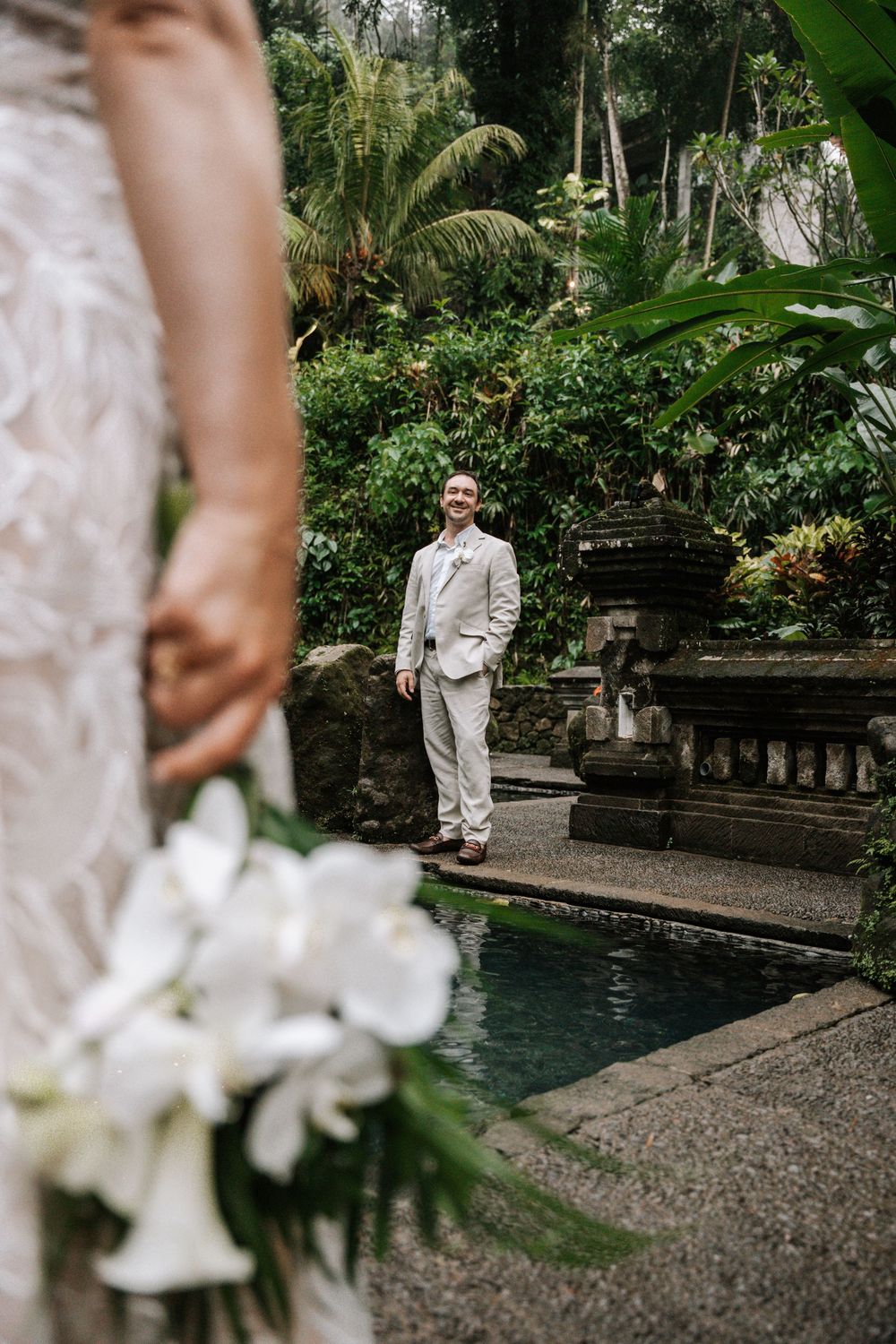 A couple in elegant white attire poses by a tropical garden pool during their destination wedding photoshoot.