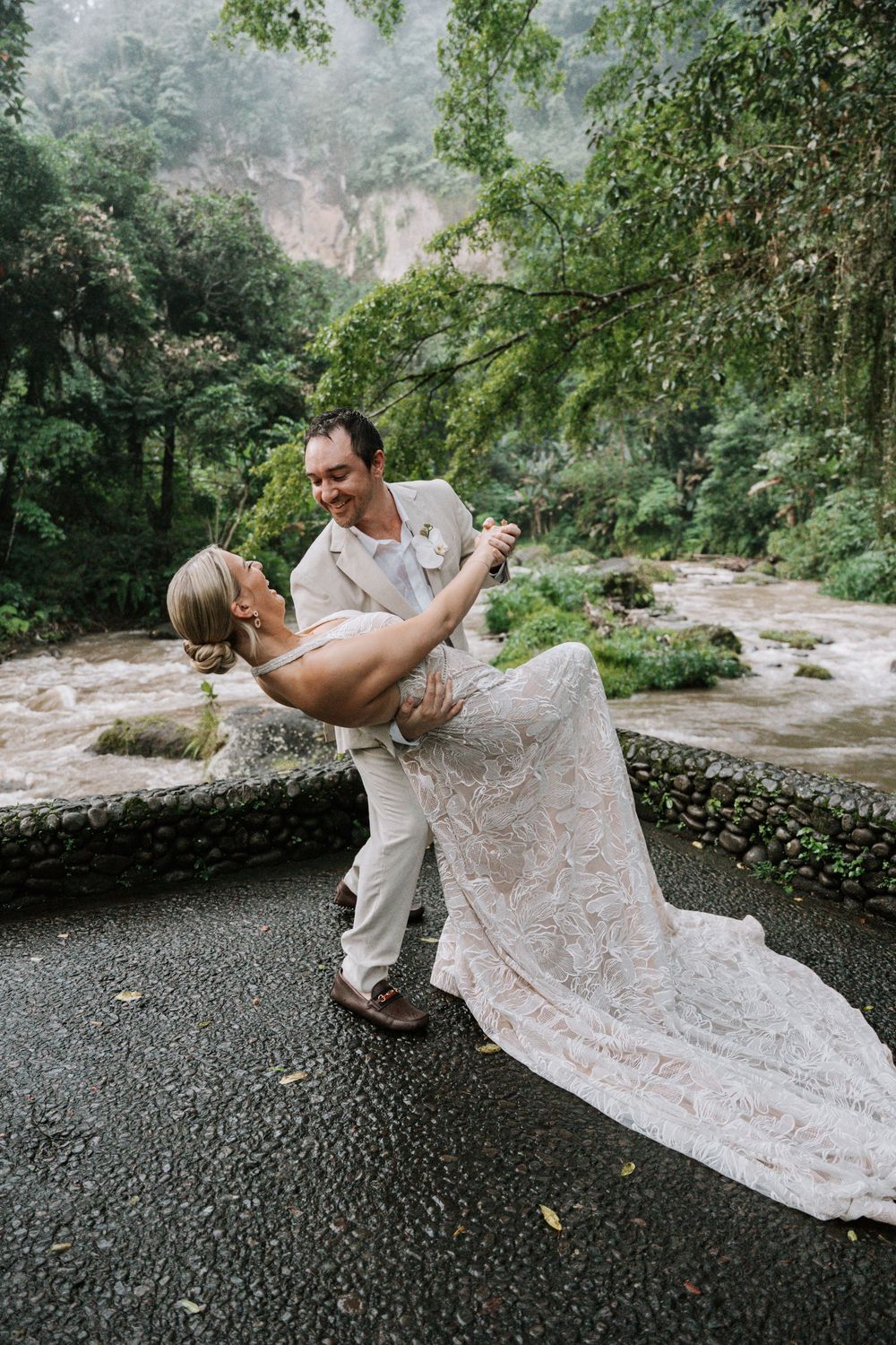 A couple in formal attire performs a dramatic dip dance move on a stone patio near a scenic mountain backdrop.