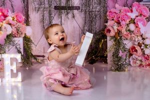 Baby in pink dress sits among floral decorations and letter props in a professional photo studio setting.