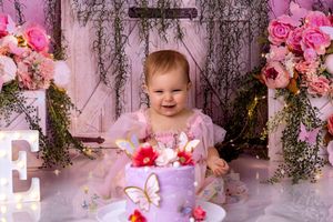 A baby in a purple tutu dress poses in a dreamy pink floral garden setting with roses and hanging vines.