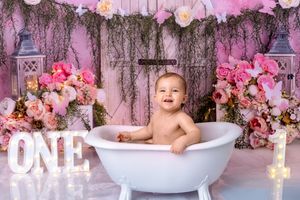Baby sitting in white bathtub surrounded by pink floral decorations and rustic wooden backdrop.