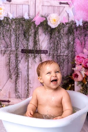 A baby sits in a white bathtub with pink floral decorations and hanging greenery in the background.