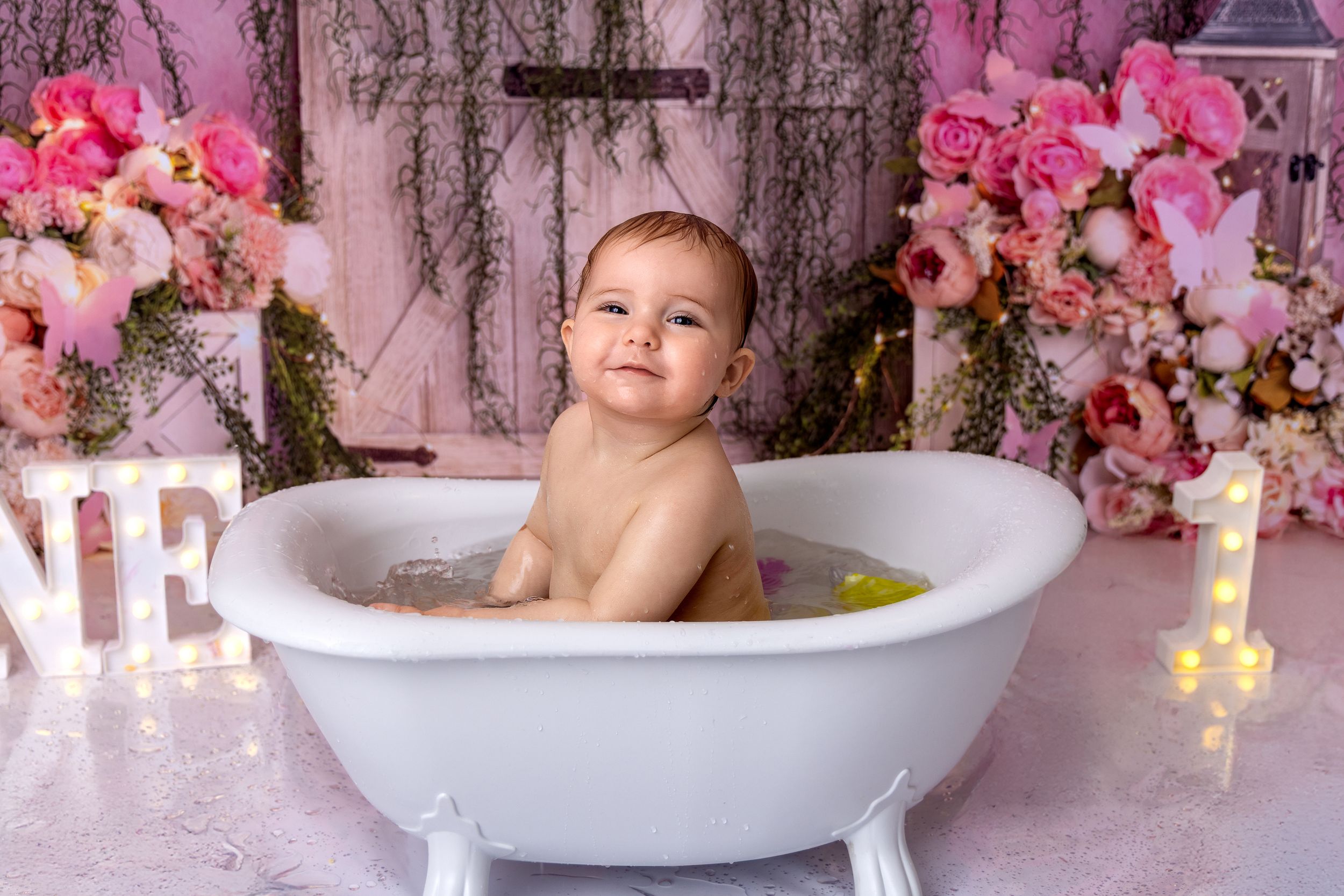 A white bathtub surrounded by pink flowers creates a dreamy setup for baby photography.