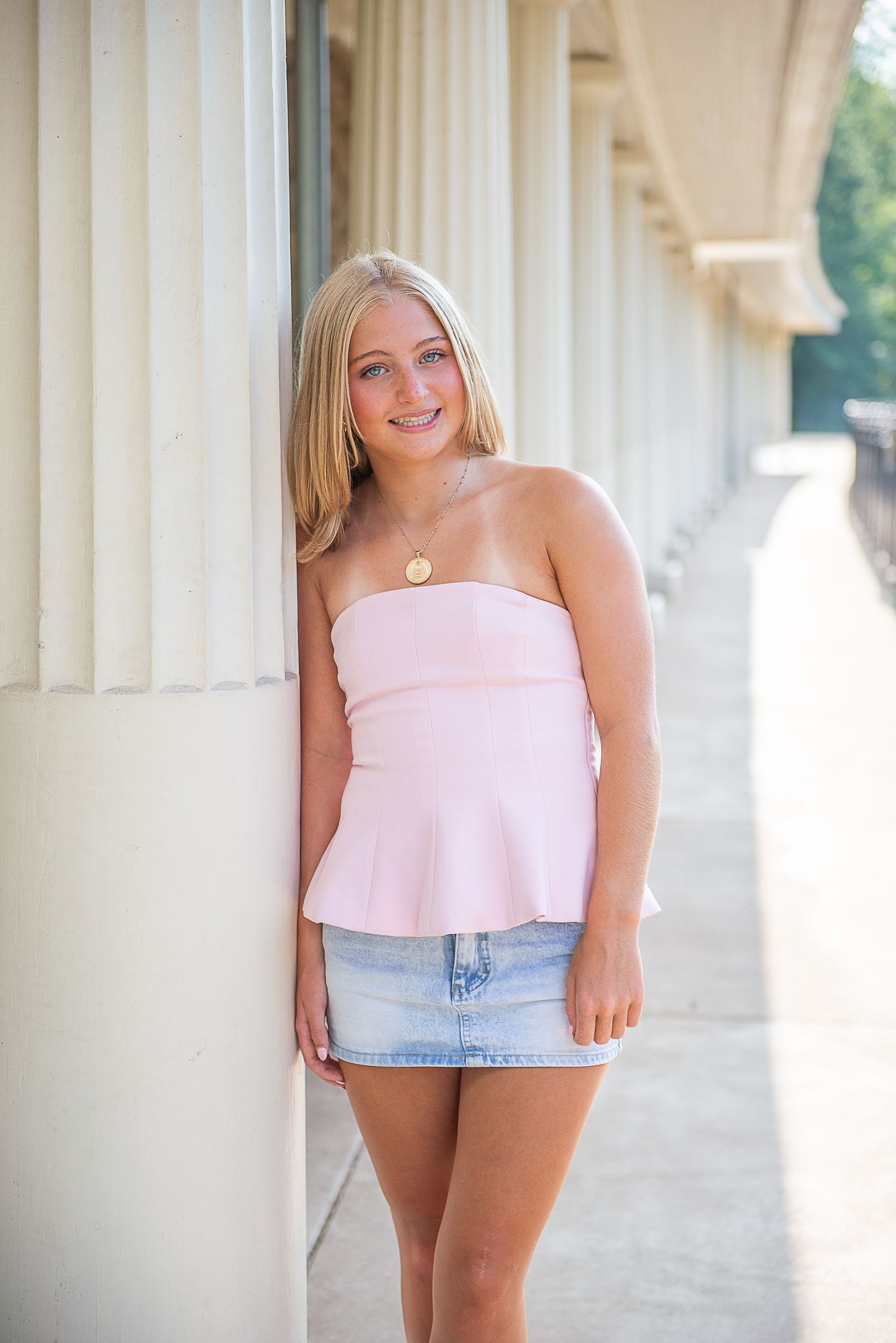 A person in a pink crop top and denim shorts poses between white columns in a summery outdoor setting.