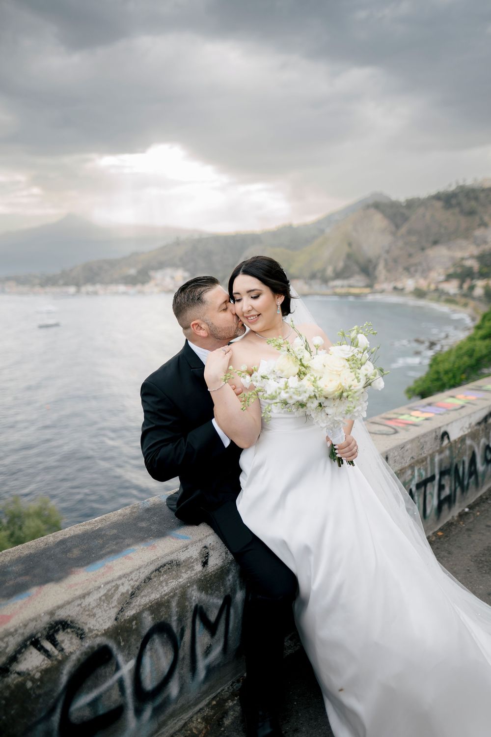 A romantic wedding couple shares a kiss along a coastal wall with mountains and ocean in the background.