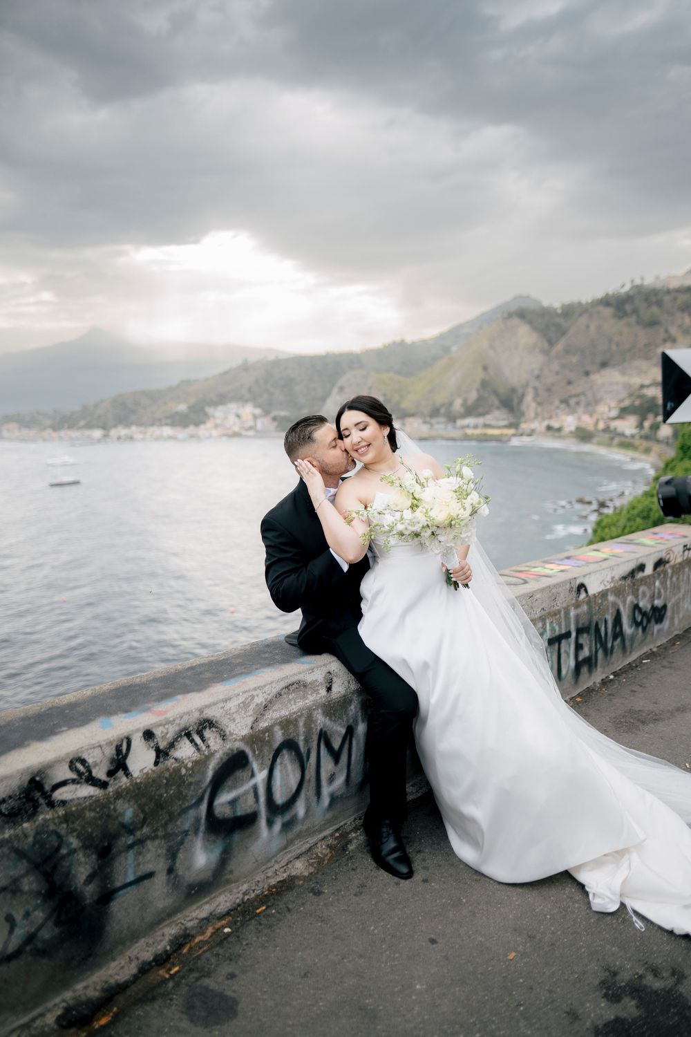 A wedding couple embraces dramatically against a cloudy coastal backdrop with graffiti wall.