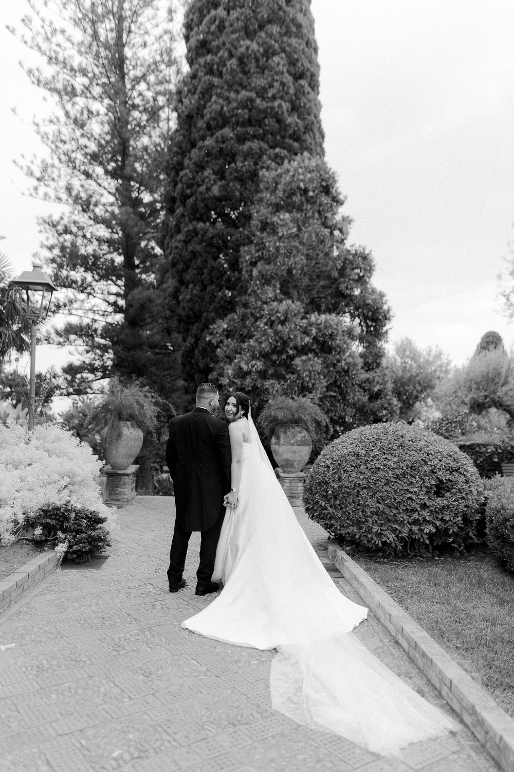A wedding couple walks along a garden path lined with evergreen trees and manicured shrubs.