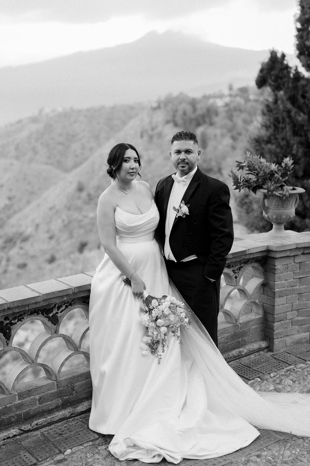 A couple in formal wedding attire share romantic moments on a stone wall overlooking mountain scenery.