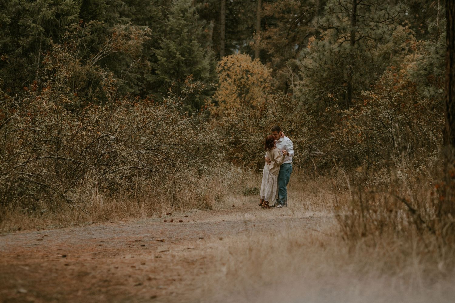 A couple stands together in a rustic outdoor setting with autumn foliage and natural lighting.