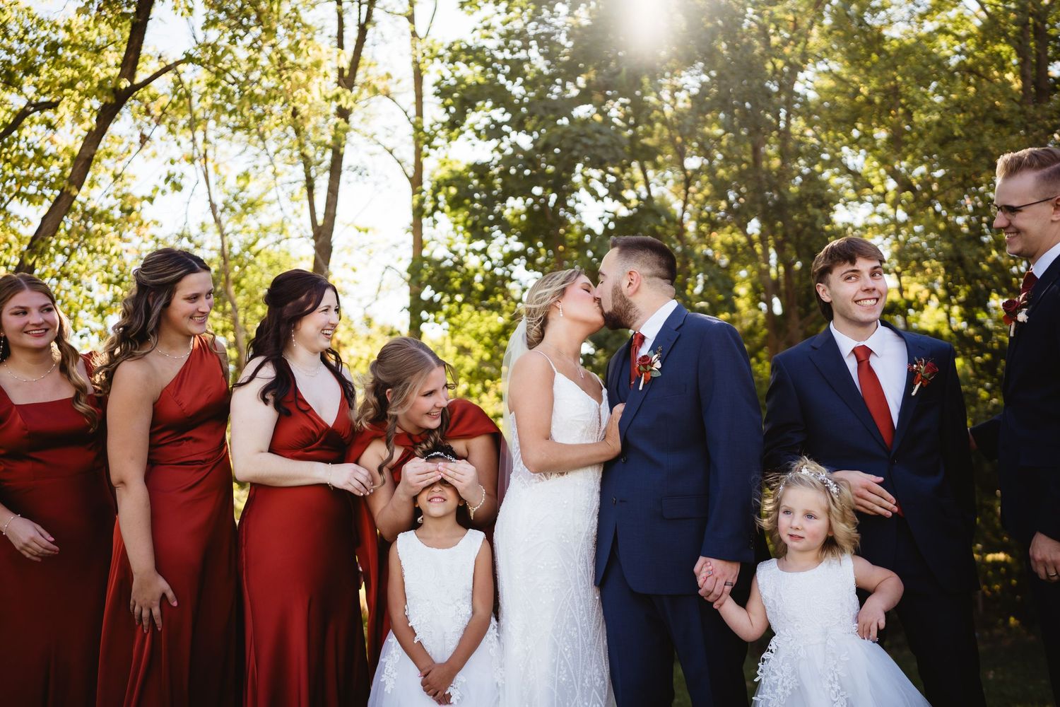 Wedding party in navy suits and burgundy dresses poses together outdoors during golden hour sunset.