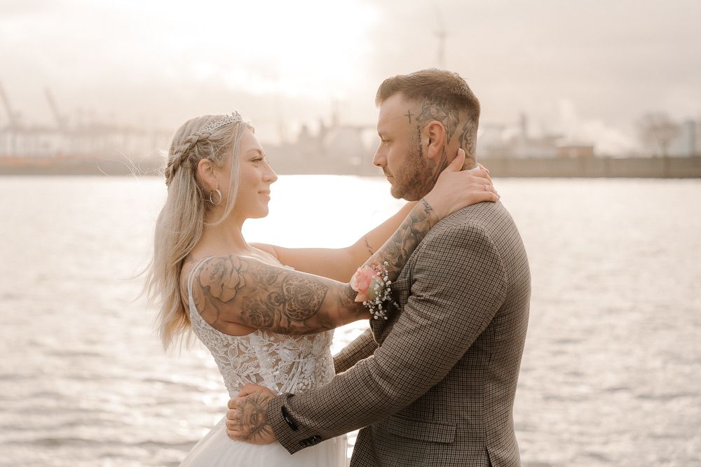 A couple embraces by the waterfront with boats and a marina visible in the background during sunset.